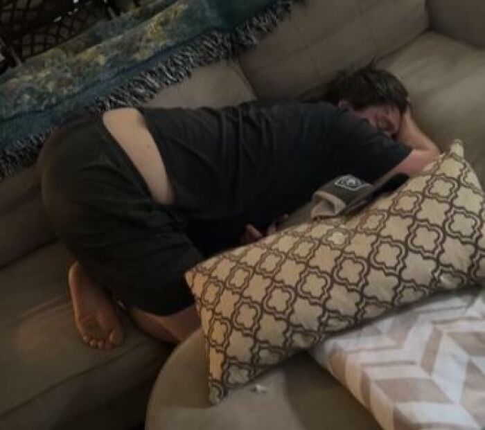 Person sleeping uncomfortably on a couch curled around pillows in one of the most hilarious and unhinged ways.