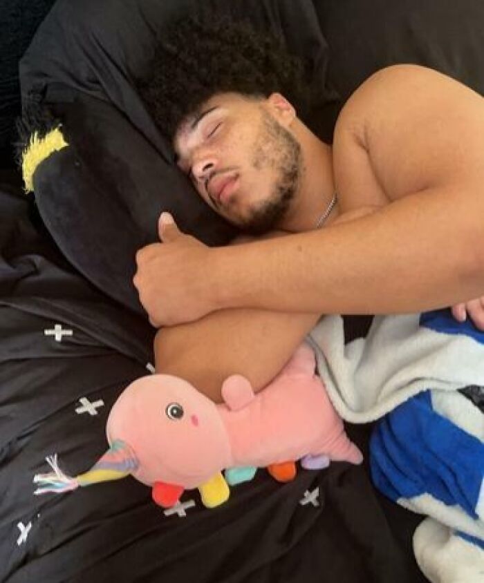 Man sleeping hugging a colorful plush toy in a funny and unhinged way on a black bed with patterned sheets.