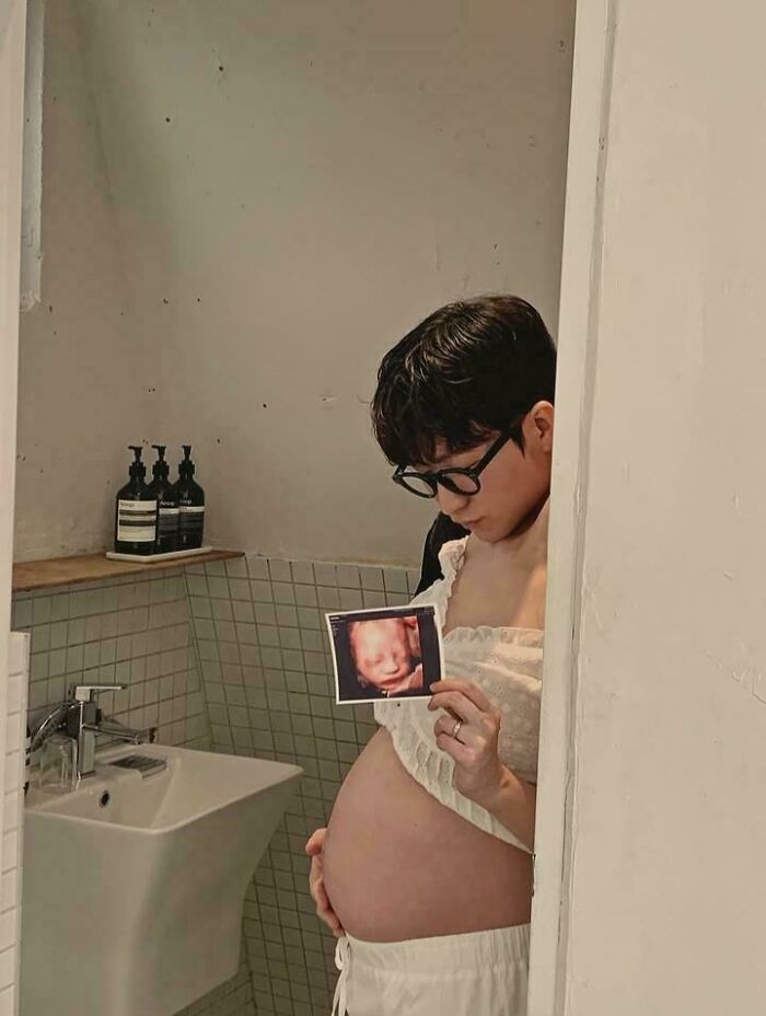 Person with glasses holding sonogram photo and showing a pregnant belly, a misleading and confusing photo.