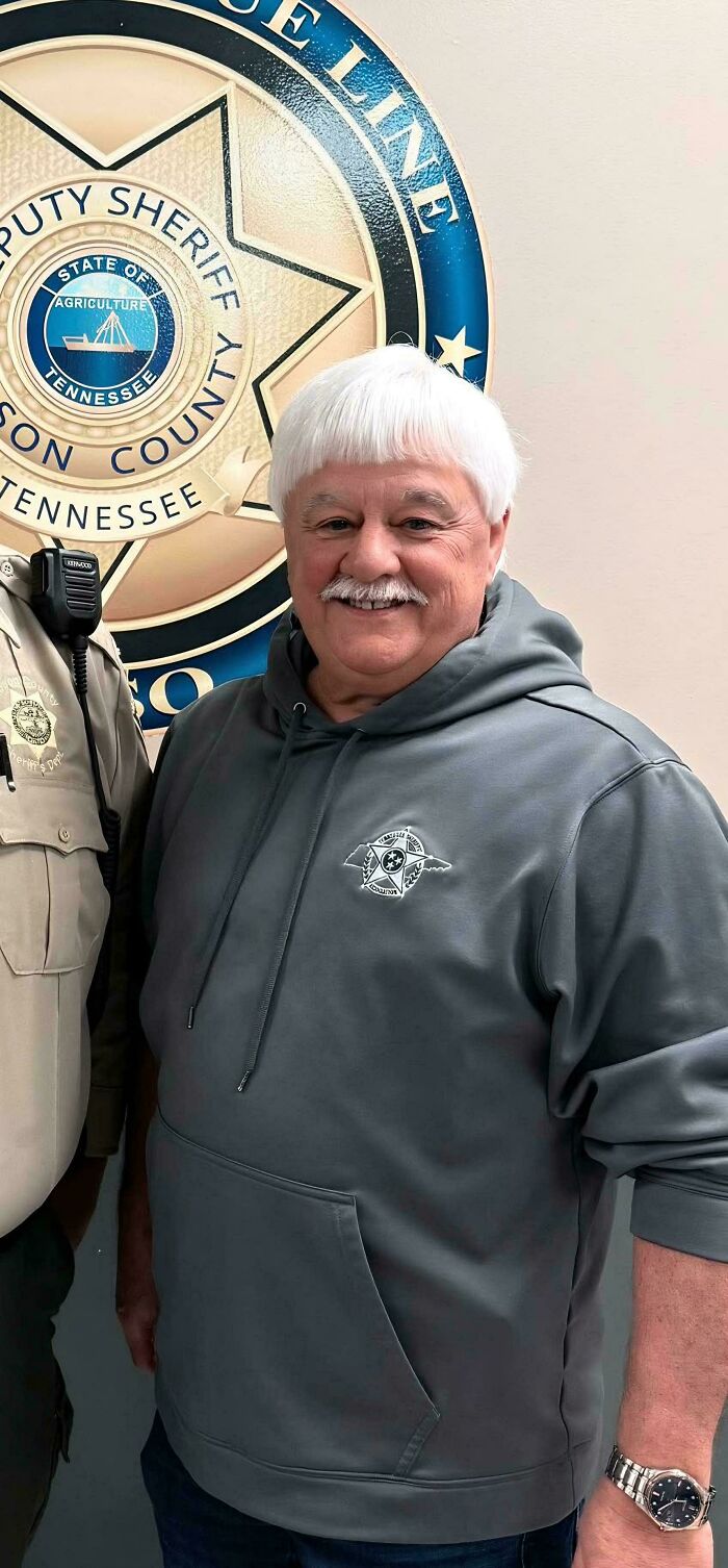 A smiling man with a white bowl cut and mustache, a bad hair mistake, wearing a gray hoodie.