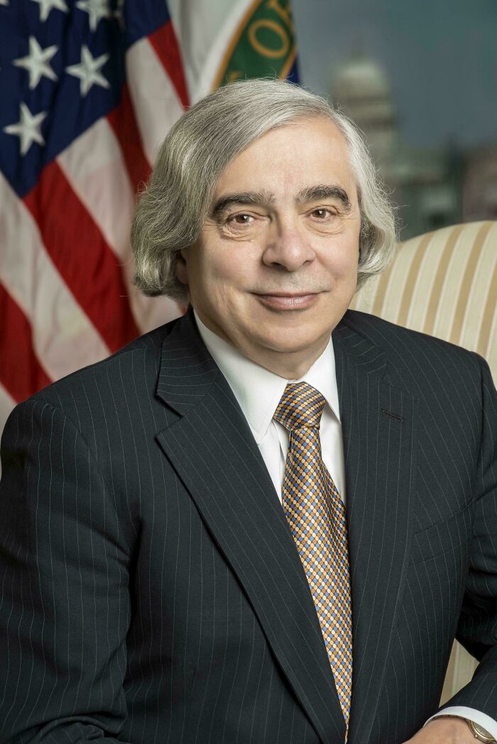 A man with a white, shaggy hairstyle, a bad hair mistake, wearing a suit in front of an American flag.