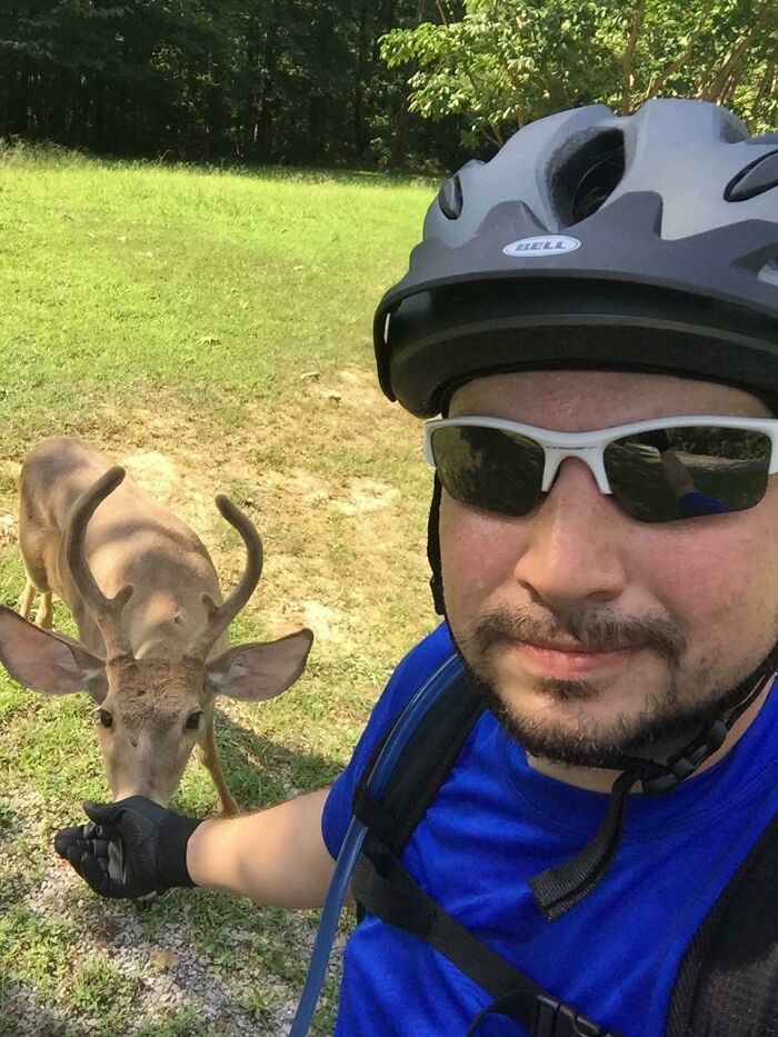 Man in a bike helmet and sunglasses having a wild and wholesome animal encounter with a young deer in a grassy area.