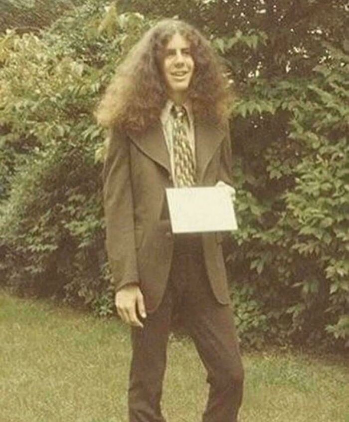 A young man with extremely voluminous, curly brown hair, wearing a suit outdoors, an example of a bad hair mistake.