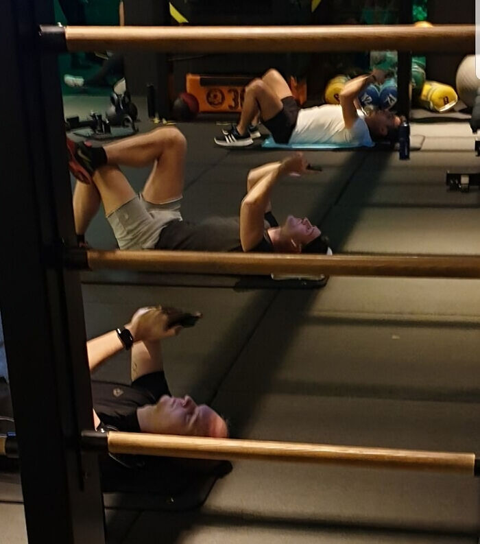 Three people lying on the gym floor using phones and medicine balls during a workout session at the gym.