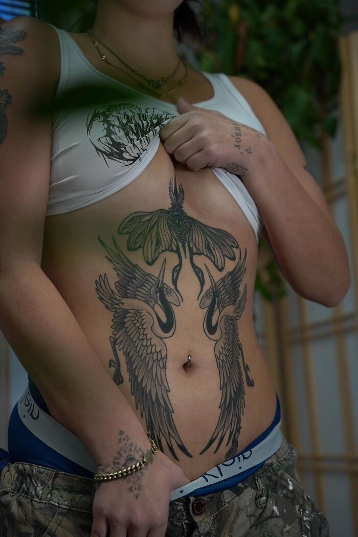 A person with a navel piercing reveals stunning tattoos of a moth and two cranes on their stomach, a happy, forever body art choice.