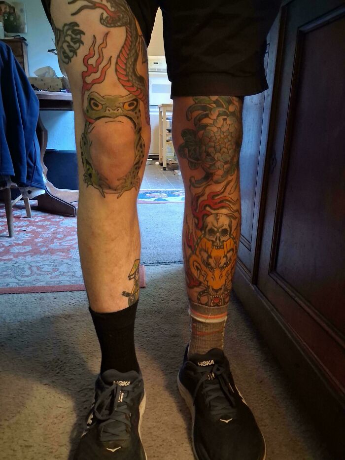 A person's legs covered in various tattoos, including a frog and a skull with flames, showcasing diverse tattoos.