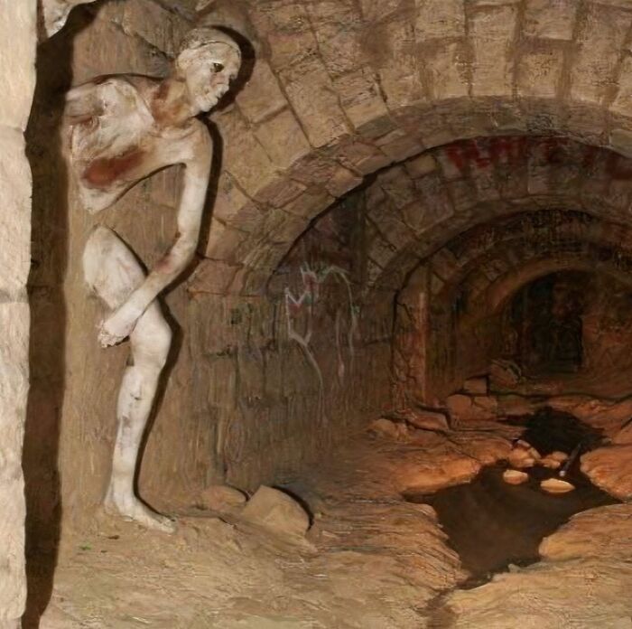 Creepy and weird photo of a decayed humanoid figure crouching inside a dark, stone-arched underground tunnel.