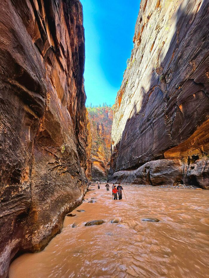 Hikers navigate a narrow canyon with steep rock walls and a flowing brown river. A great outdoor activity for dads.