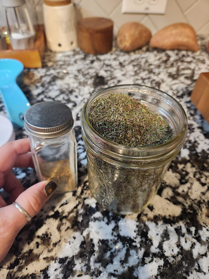 A hand holds a spice shaker next to a jar of dried herbs on a granite counter, showing frugal tips for a new world.
