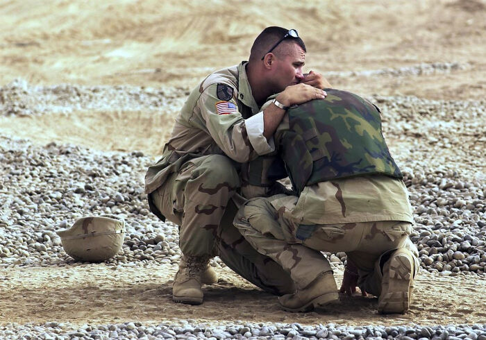 A historical photo of two soldiers in camouflage embracing on a rocky, sandy ground, one with an American flag patch.