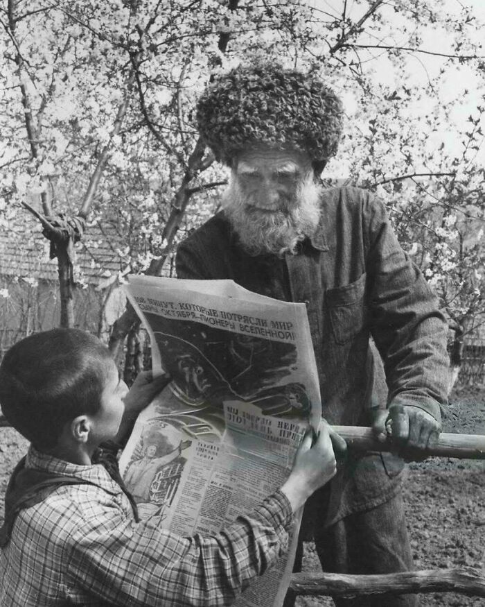 A black and white historical photo of a boy and an older man with a beard and fur hat reading a newspaper together.