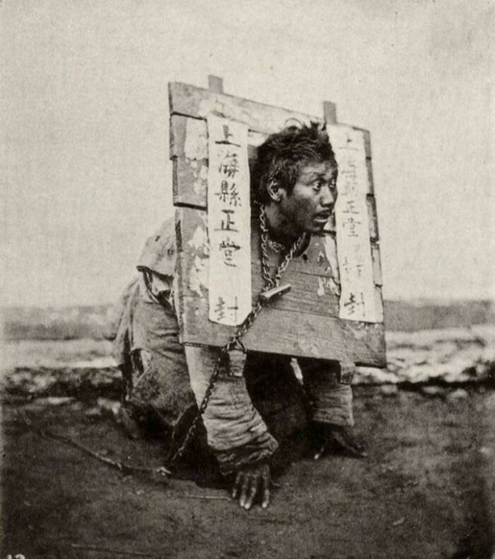 A historical photo of a man in stocks, kneeling with a wooden frame around his neck and chained, on a dirt ground.