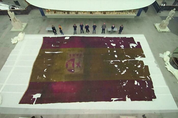 Overhead view of a large, tattered historical Spanish flag laid out, surrounded by people, showcasing historical photos.