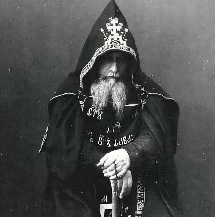 A black and white portrait of a man with a long beard wearing a dark, elaborate hooded robe, from historical photos.