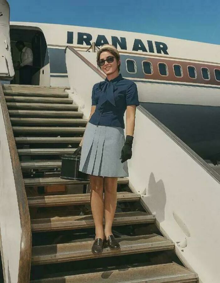 A flight attendant in uniform smiling on the stairs of an Iran Air plane, capturing historical photos of travel.