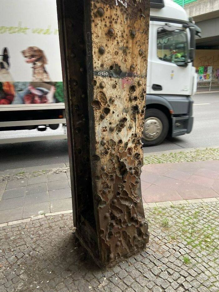 A heavily corroded metal post on a sidewalk next to a road with a truck passing, showing historical photos of decay.