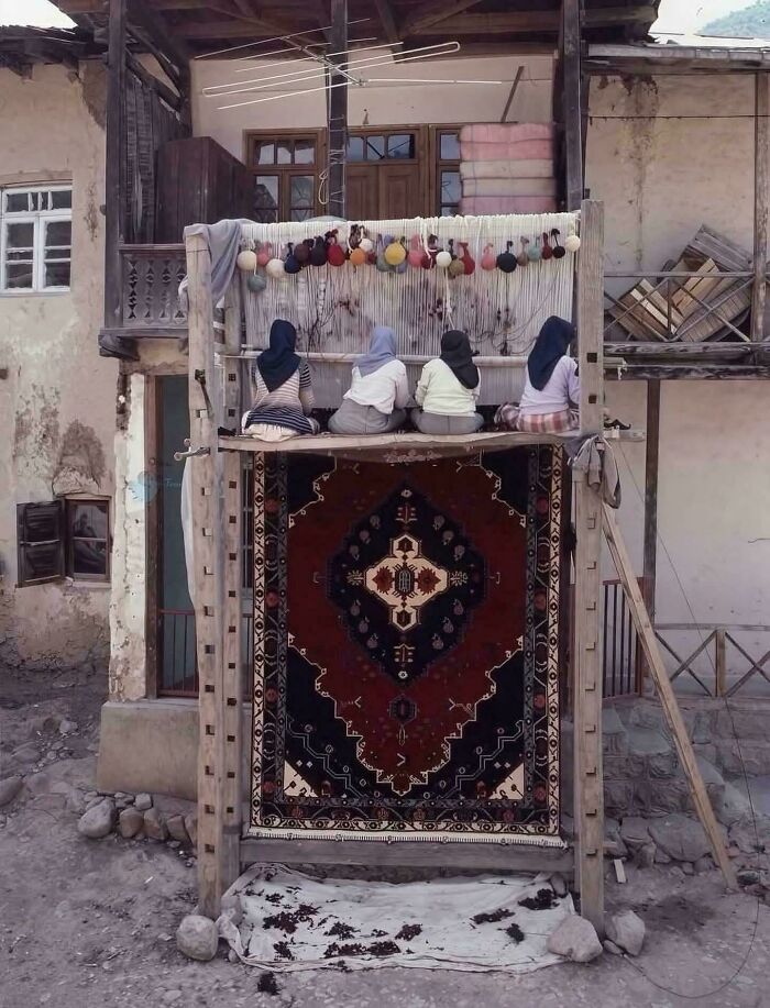 A historical photo showing four women weaving an intricate, large rug on a loom outside a traditional building.