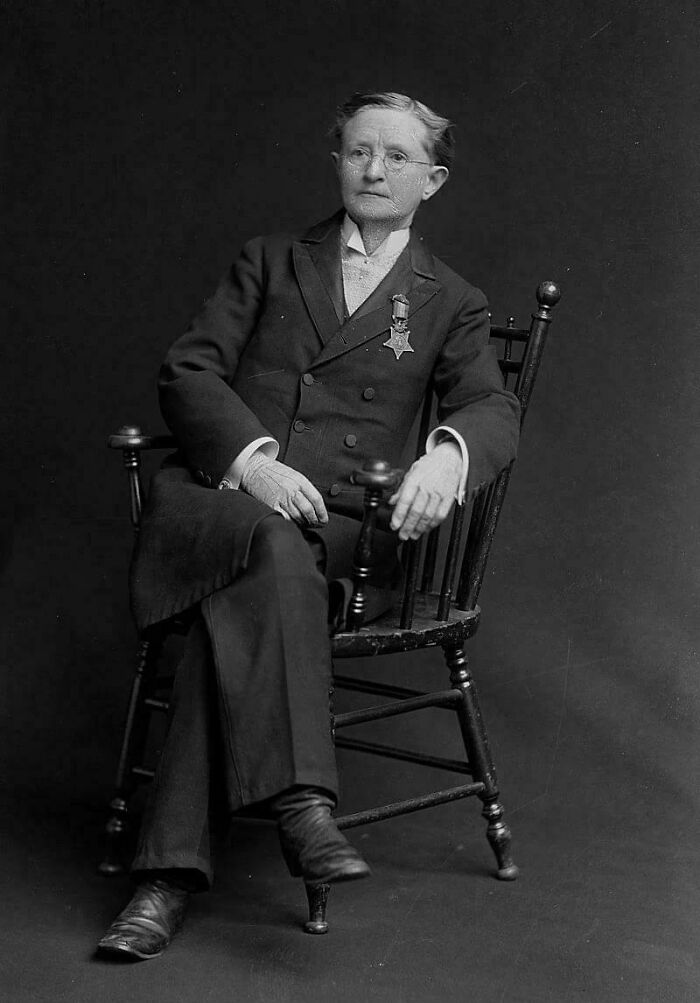 A historical photo of Dr. Mary Edwards Walker, a woman in a suit with medals, seated in a wooden chair.
