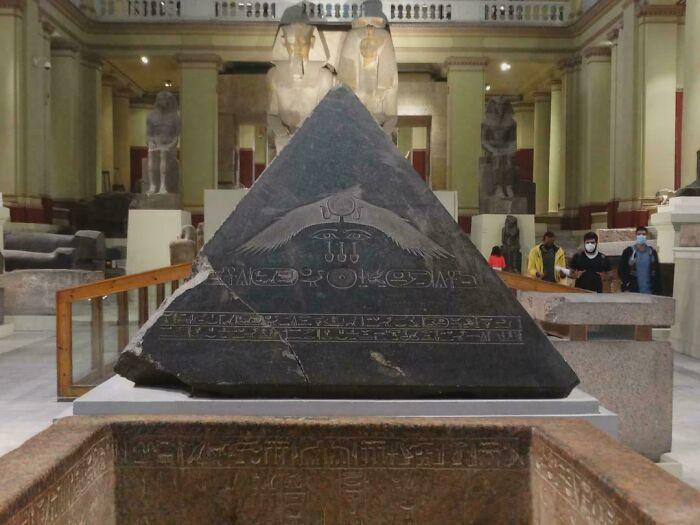 A historical photo of a large black pyramid-shaped stone with hieroglyphs in a museum, surrounded by statues.