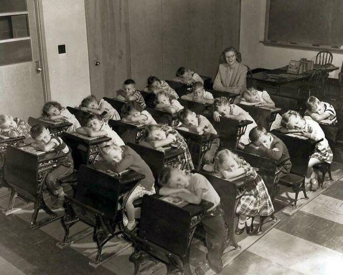 A historical photo of a classroom of young children napping at their desks, with a teacher watching.