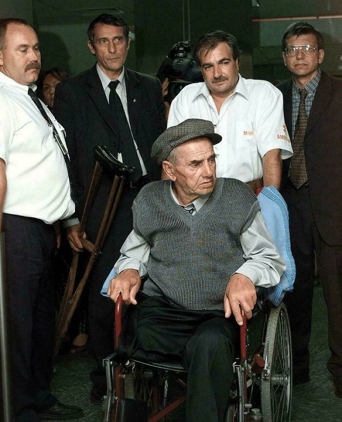 A historical photo of an older man in a cap and vest in a wheelchair, surrounded by men in suits.