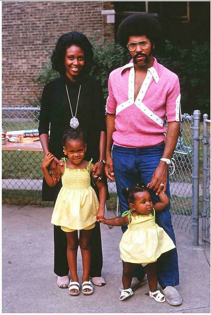 A candid old photo of a family of four, parents and two young daughters, standing outdoors in casual attire.