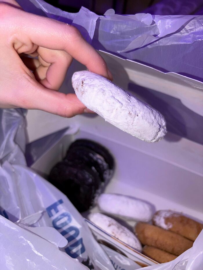 Hand holding powdered doughnut with box of assorted doughnuts inside bag