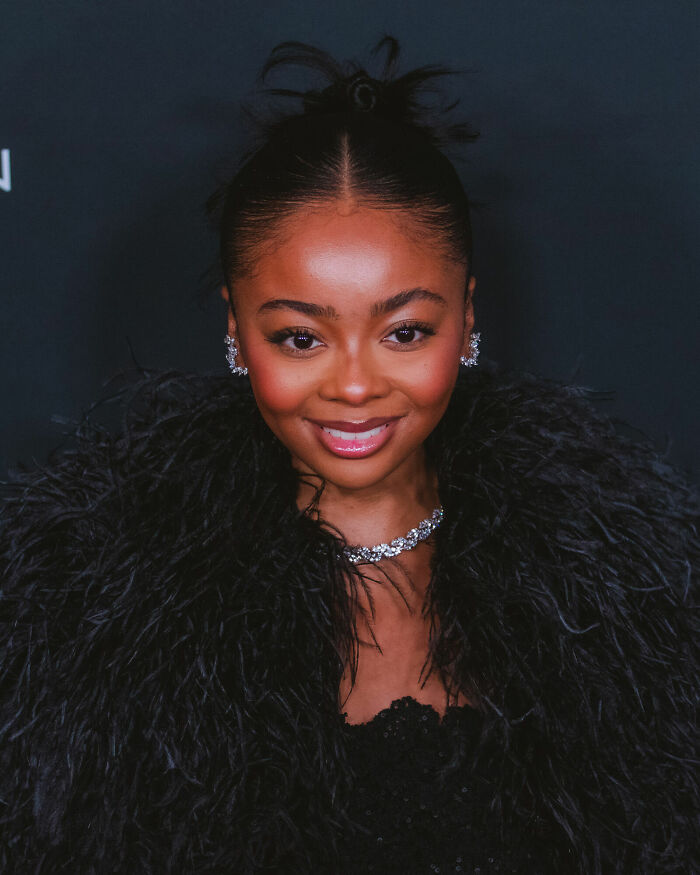 Skai Jackson smiling at an event, wearing a black feathered outfit and diamond jewelry for a formal occasion.