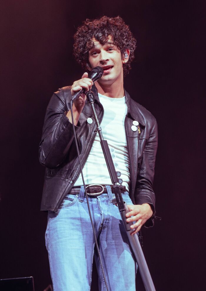Matty Healy performing live on stage, wearing a leather jacket and holding a microphone during a concert.