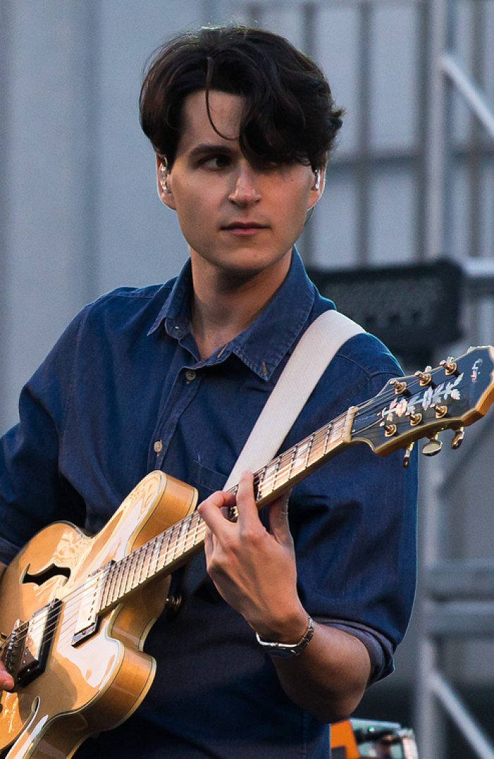 Ezra Koenig playing guitar on stage, wearing a blue shirt and focused during a live musical performance.