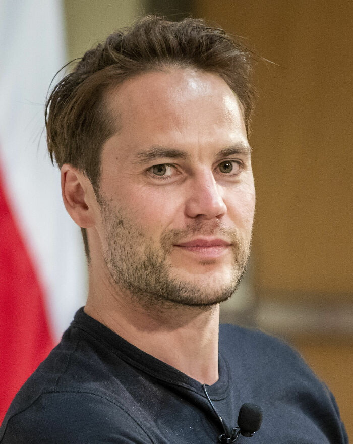 Taylor Kitsch with short hair and facial stubble, wearing a black shirt during a public speaking event.