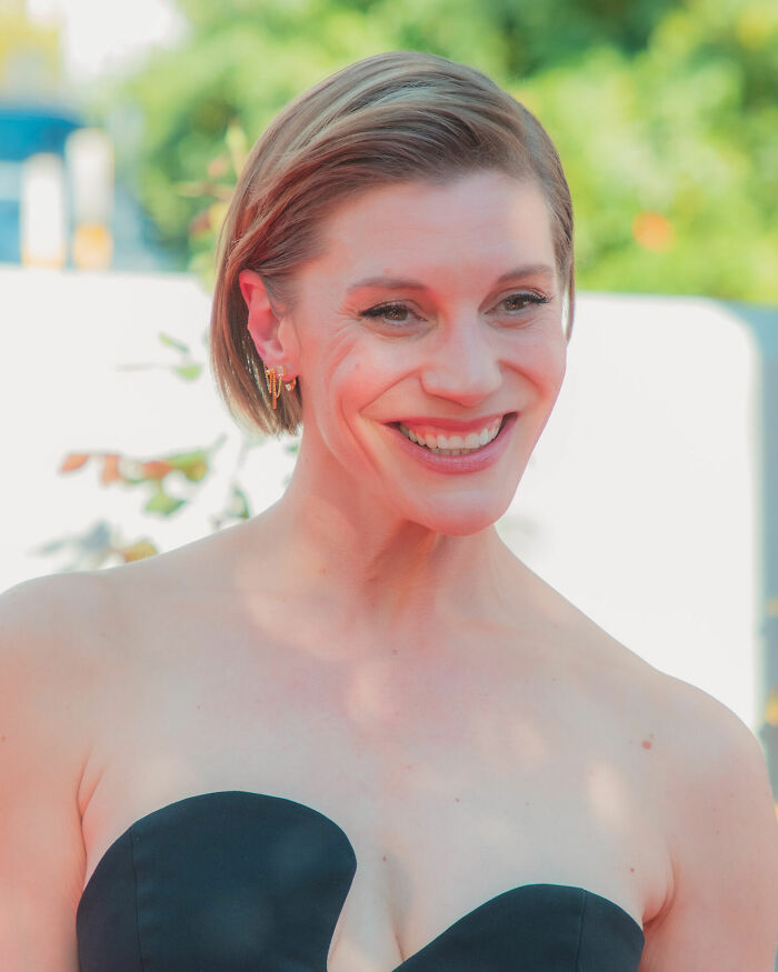 Katee Sackhoff smiling at an outdoor event wearing a strapless black dress with natural makeup and short hair.