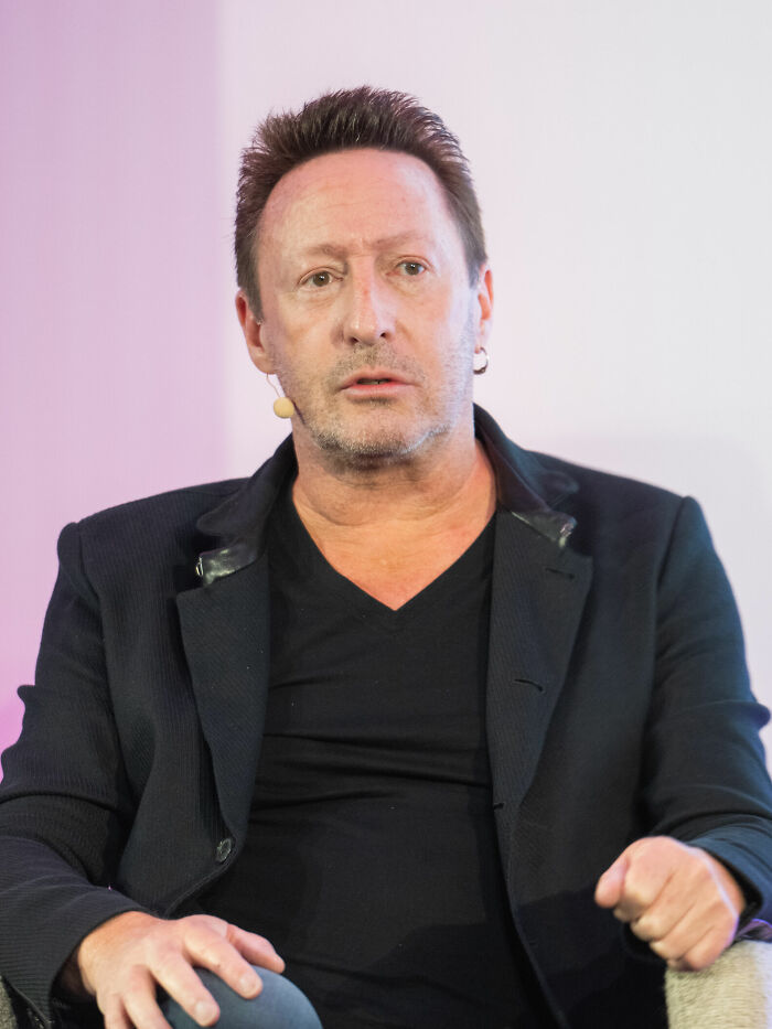 Julian Lennon speaking at an event, wearing a black jacket and microphone headset, sharing bio and career highlights.