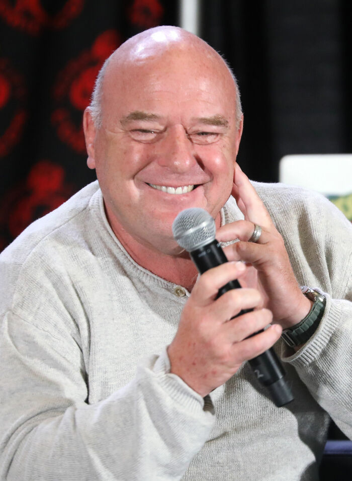 Dean Norris smiling while holding a microphone, wearing a light sweater, participating in a casual speaking event.