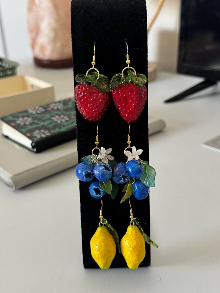 Three pairs of handmade projects fruit-themed earrings: red strawberries, blue blueberries with white flowers, and yellow lemons, hanging on a black display stand.