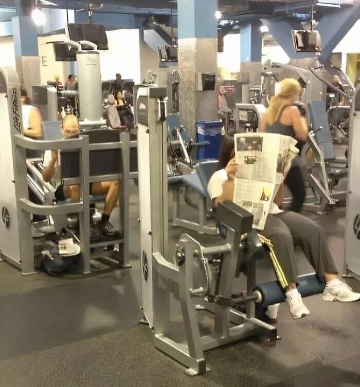 Gym scene with people using workout machines, including one person reading a newspaper, unusual gym behavior caught.