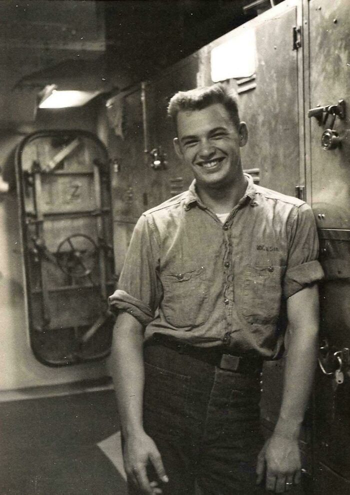 A candid old photo of a smiling man in a work shirt and pants, standing inside what appears to be a ship.