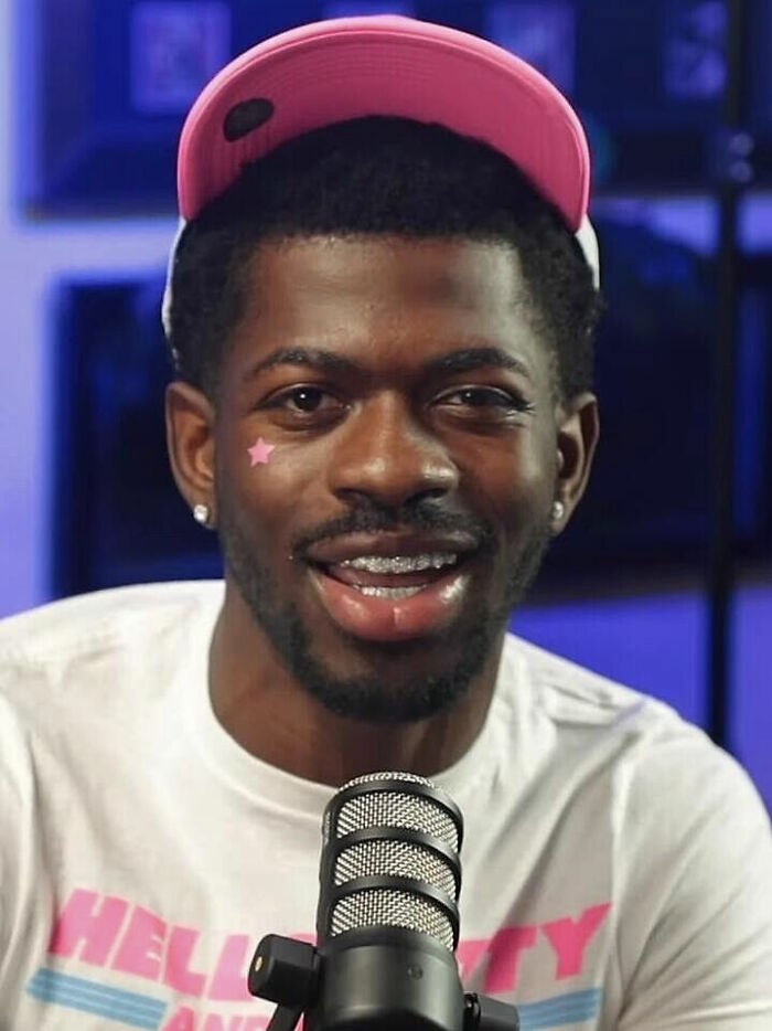 Lil Nas X wearing a pink cap and white shirt, smiling while speaking into a microphone in a studio setting.