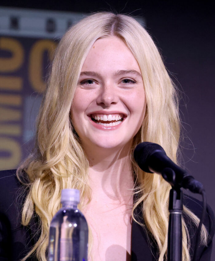 Elle Fanning speaking at a panel event, smiling with long blonde hair and a microphone in front of her.