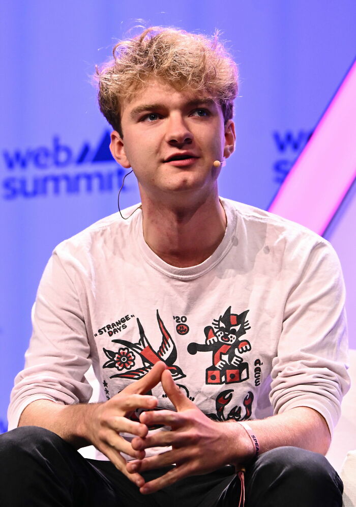 TommyInnit speaking at a conference, wearing a white graphic shirt and a headset microphone, seated on stage.