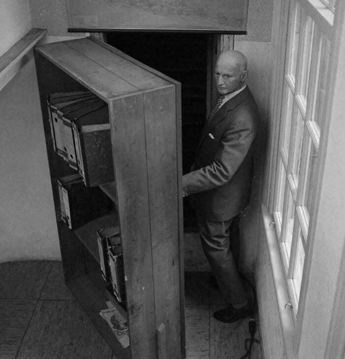 Black and white historic photo of a man in a suit opening a hidden bookshelf door in a narrow hallway.