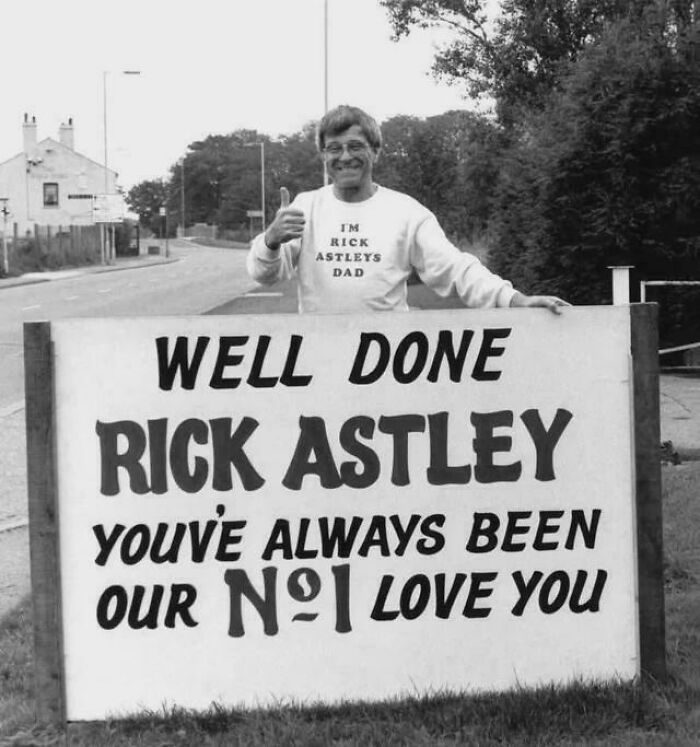 Man wearing I'm Rick Astley's Dad shirt giving thumbs up beside a historic photo with a congratulatory sign.