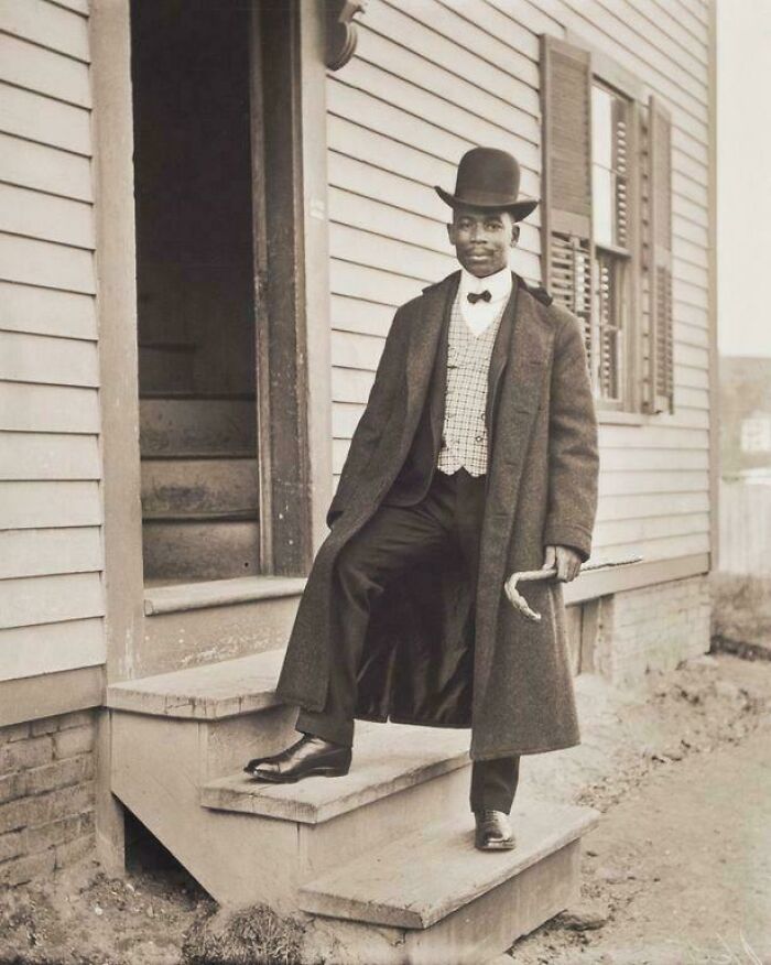 Historic photo of a stylish man in a long coat and top hat standing on wooden steps, showcasing life in a different era.