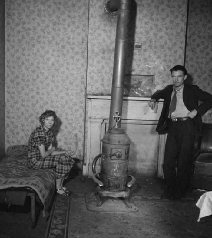 Young woman sitting on a bed and man standing by a stove in a historic photo showing life in a different era.