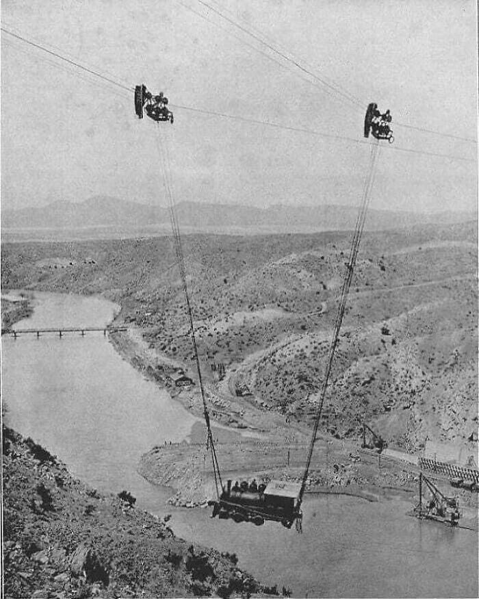 Historic photo showing early 20th-century cable car transport over a river in a rugged landscape.