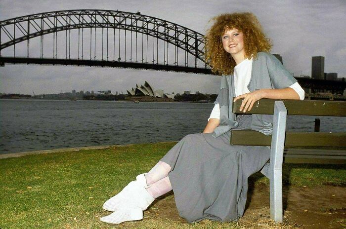 Young woman with curly hair sitting on a bench near Sydney Harbour Bridge, a rare and fascinating historic photo.