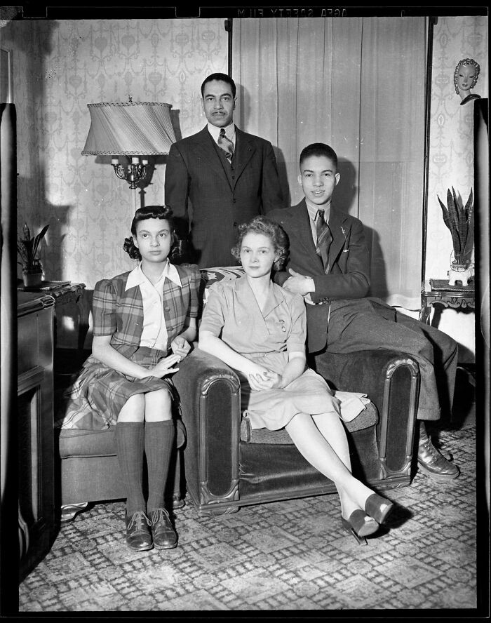 Historic photo of a family dressed in vintage clothing sitting and standing in a living room from a different era.