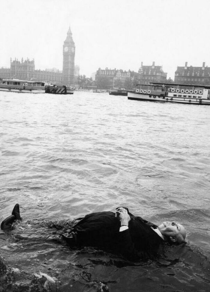 Man in a suit floating in a river with historic London landmarks in the background, rare historic photo capturing different era life.