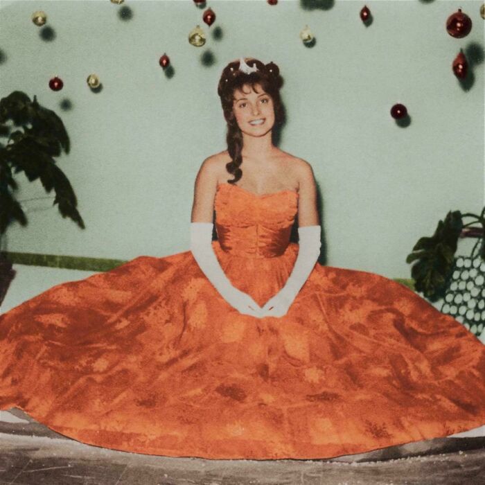 Young woman in a vintage orange gown with white gloves, posing among hanging ornaments in a historic photo.