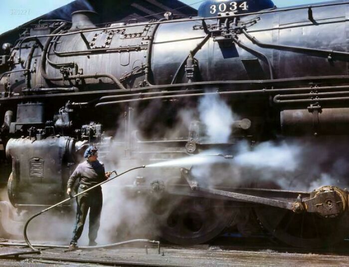 Historic photo of a worker cleaning a large steam locomotive surrounded by steam and smoke from a different era.